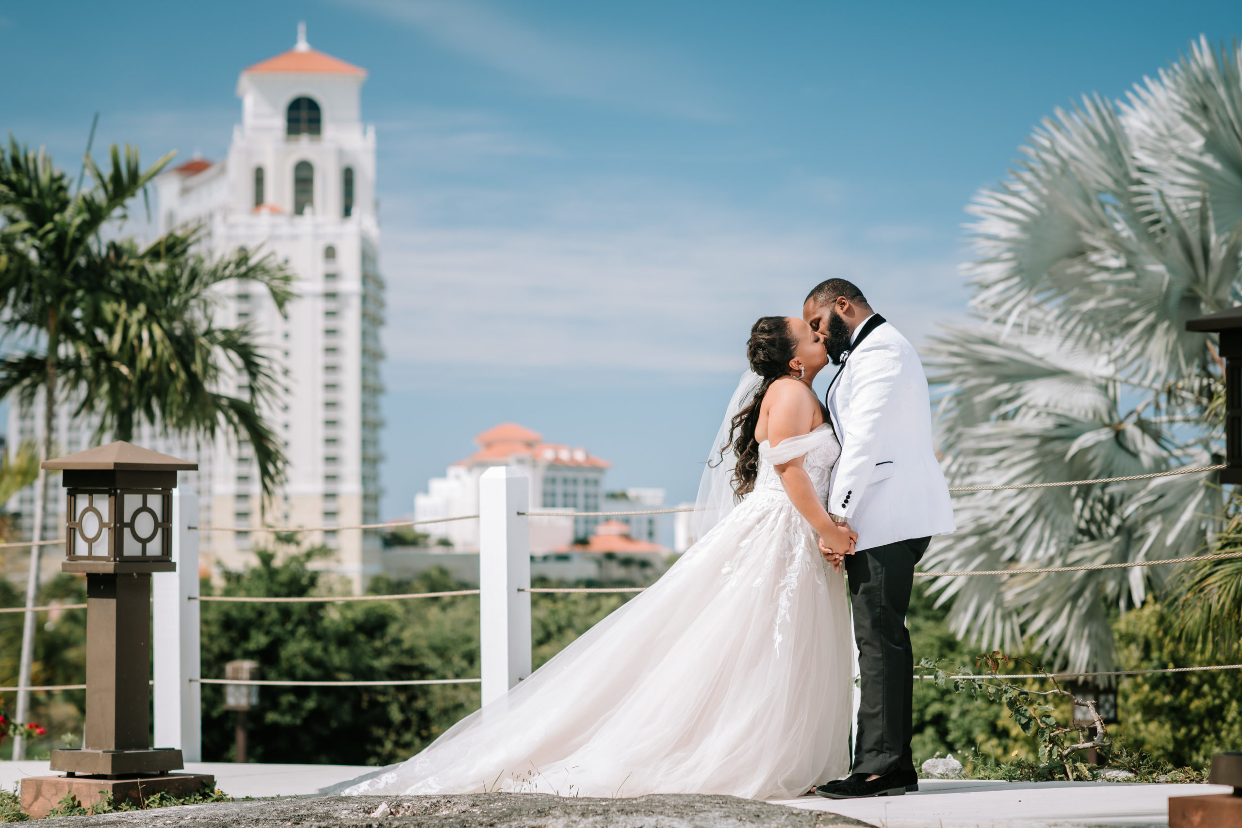 bride kissing room in front of a beautiful blue sky and large white hotel building in the background in the bahamas